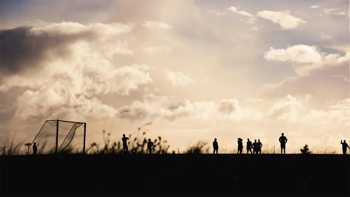 Silhouetten von Menschen und einem Fußballtor auf einem Hügel vor dramatischem Wolkenhimmel