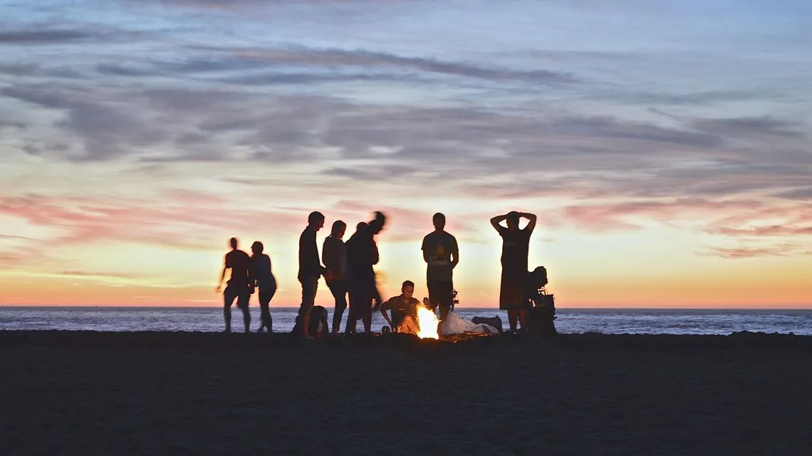 Gruppe von Menschen steht und sitzt um ein Lagerfeuer am Strand bei Sonnenuntergang