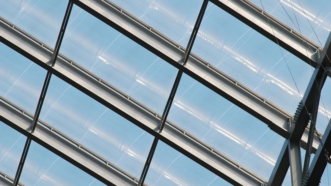 Nahaufnahme einer verglasten Dachkonstruktion mit diagonalen Stahlträgern vor blauem Himmel
