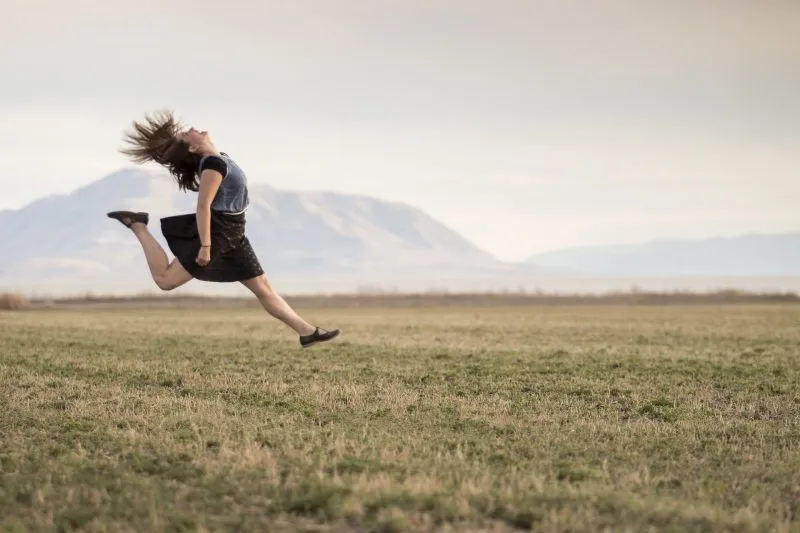 Frau springt mit wehenden Haaren über eine weite Wiese, im Hintergrund Berge am Horizont