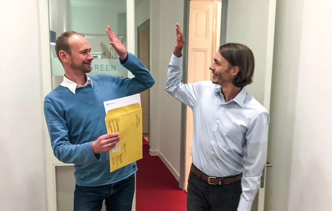 Zwei Männer geben sich im Flur ein High Five, einer hält einen gelben Umschlag in der Hand