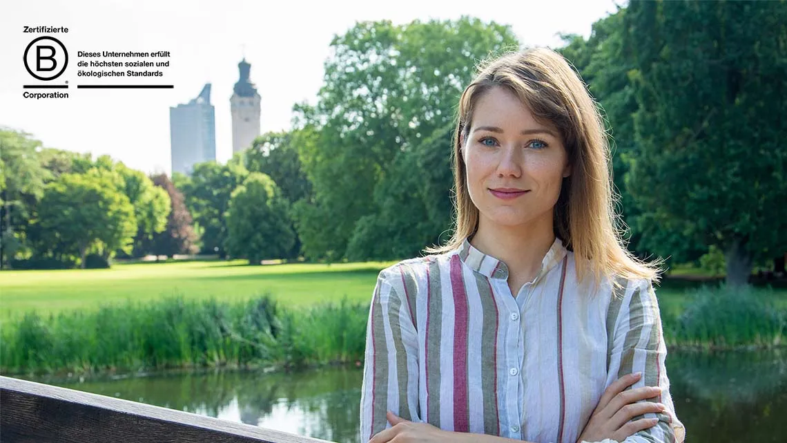 Frau mit verschränkten Armen im Park, daneben das Logo „Zertifizierte B Corporation“