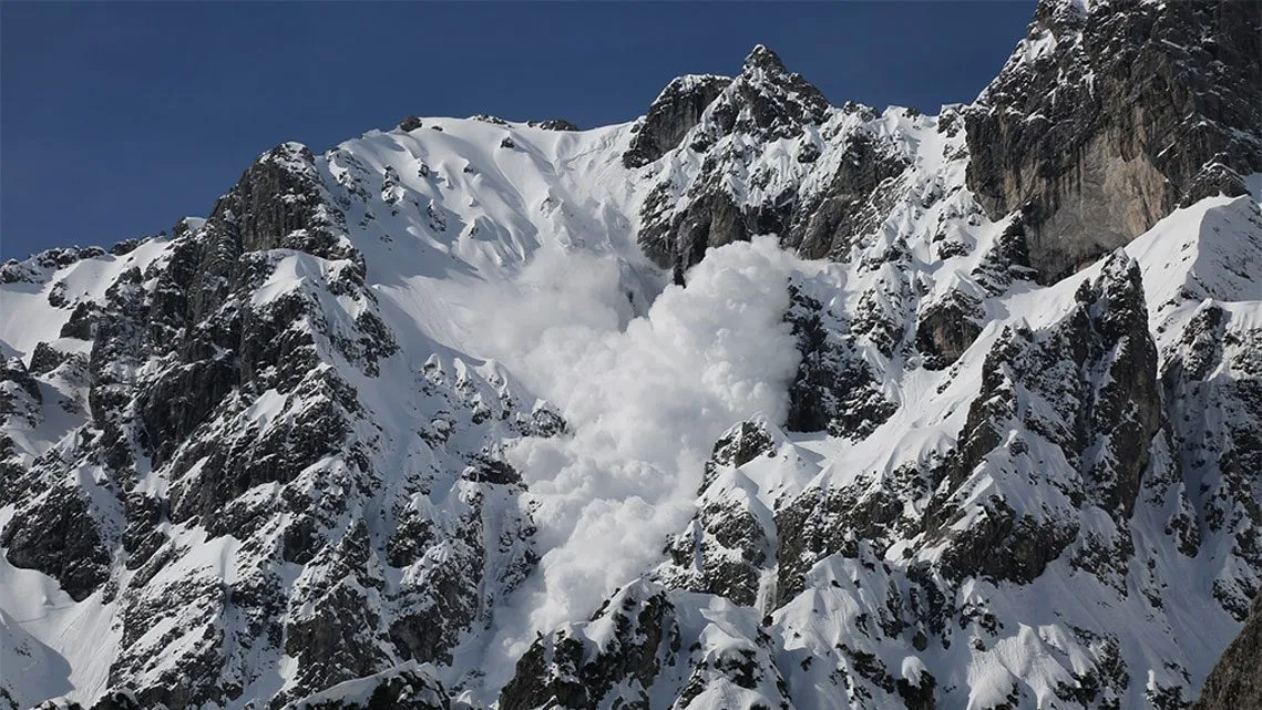 Eine Lawine bricht von einem schneebedeckten Gebirgshang ab und wirbelt Schnee in die Luft.