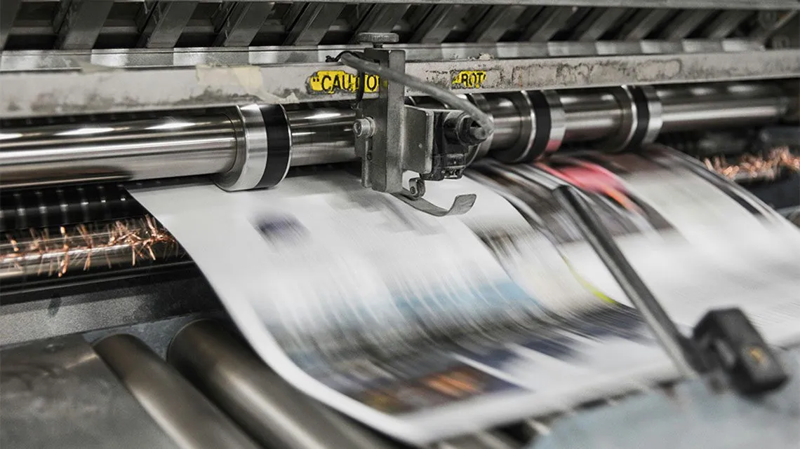 Nahaufnahme einer Druckmaschine, durch die eine bedruckte Zeitungsbahn läuft