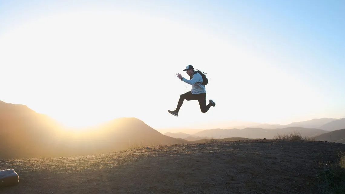 Person springt mit erhobenen Armen auf einem Hügelkamm vor Sonnenuntergang und Bergpanorama