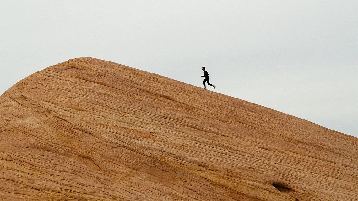 Silhouette einer joggenden Person auf dem Kamm einer Sandsteindüne vor bewölktem Himmel