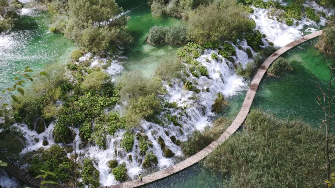 Luftaufnahme von türkisen Wasserfällen und Seen mit hölzernem Bohlenweg hindurch, umgeben von Grün