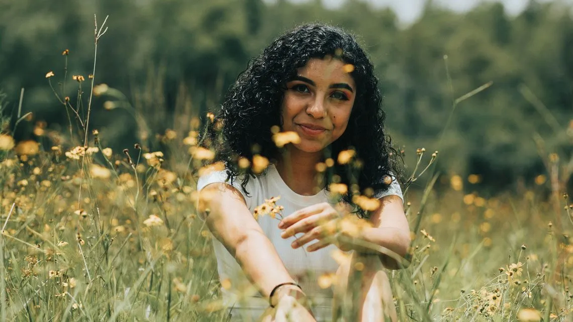 Frau mit lockigem Haar sitzt lächelnd in einer Blumenwiese mit gelben Wildblumen