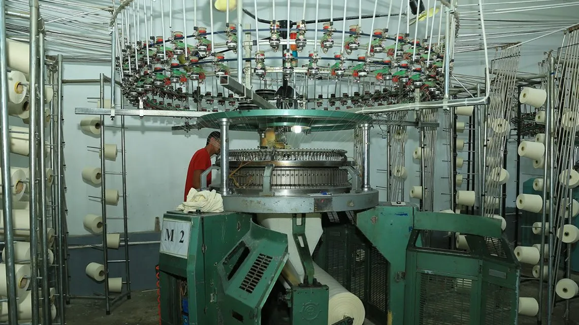 Arbeiter an einer Rundstrickmaschine in einer Textilfabrik mit zahlreichen Garnspulen