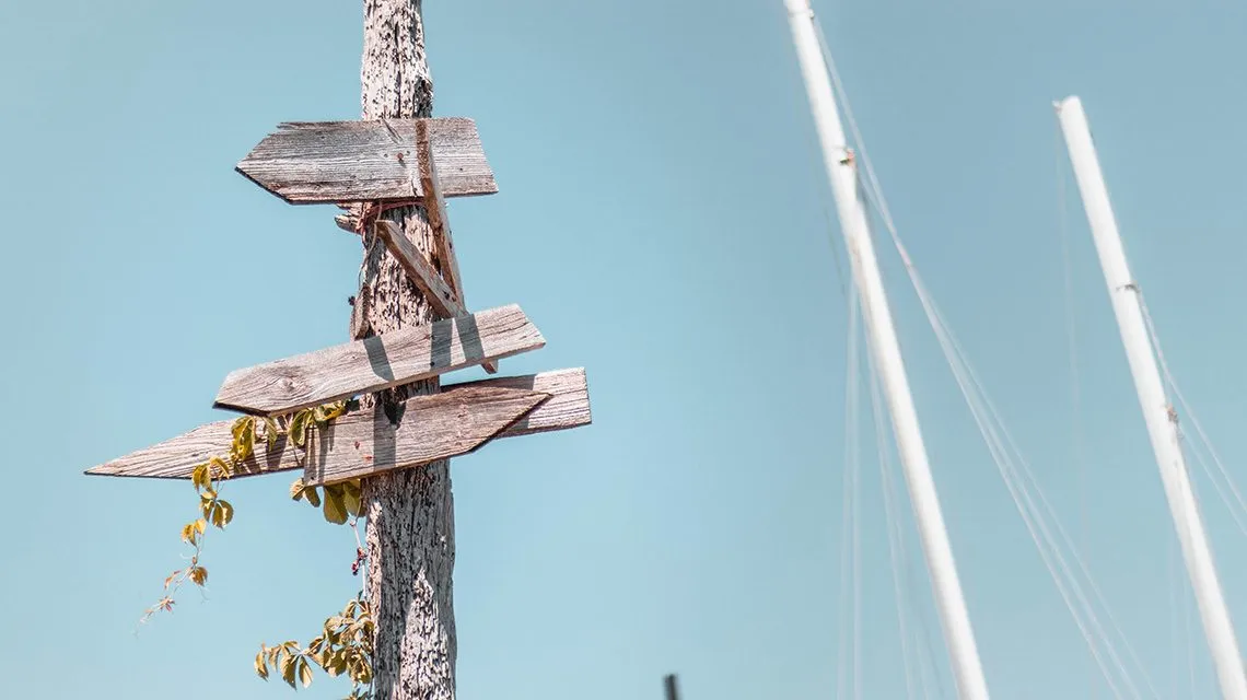Verwitterter Wegweiser mit leeren Schildern vor Segelmasten und blauem Himmel