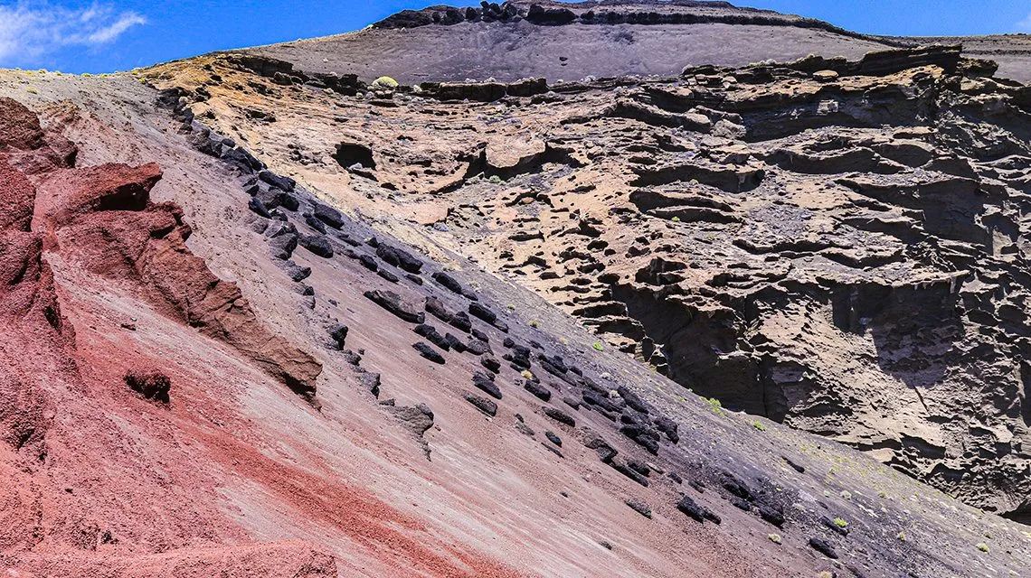 Rötlich-braune Vulkanlandschaft mit erodierten Gesteinsschichten und blauem Himmel