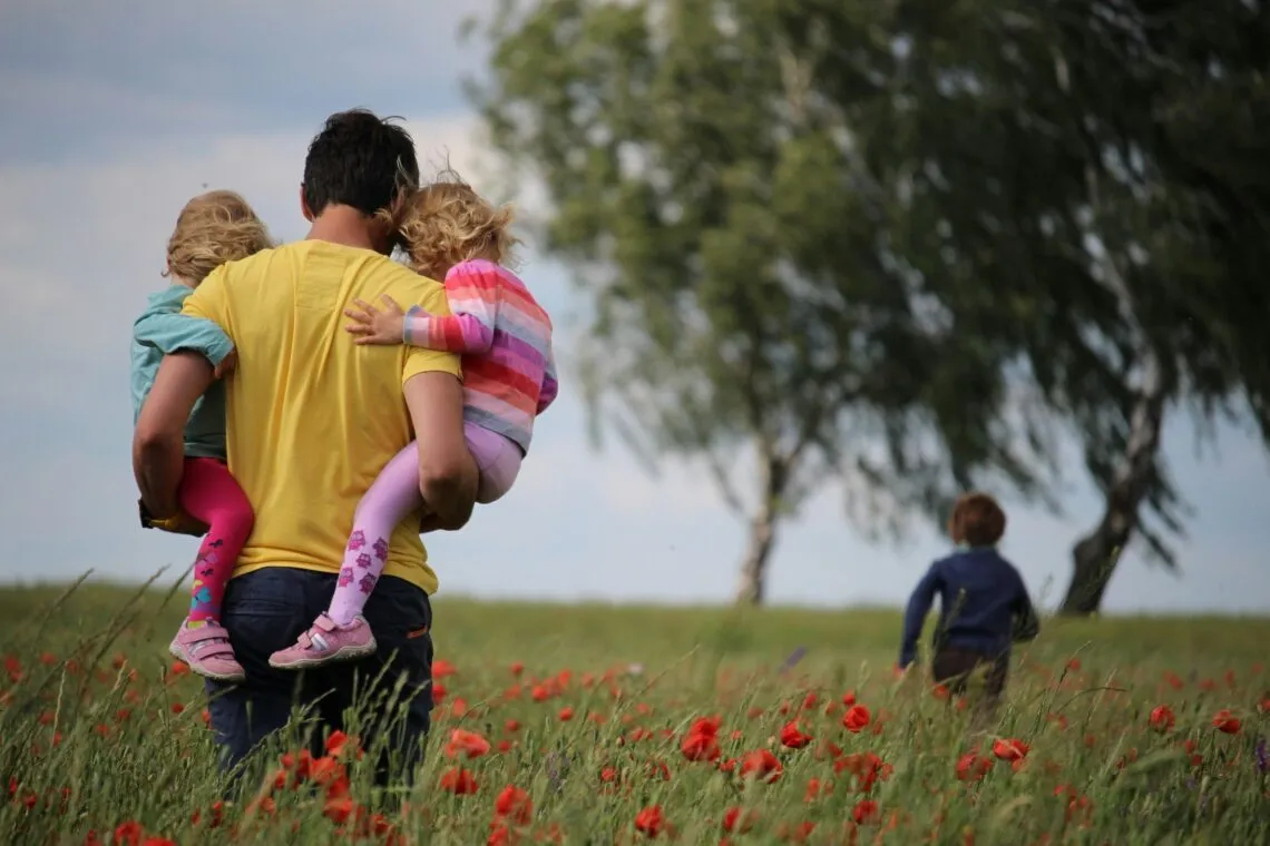 Ein Mann trägt zwei kleine Kinder auf den Armen durch ein Mohnblumenfeld, im Hintergrund läuft ein drittes Kind
