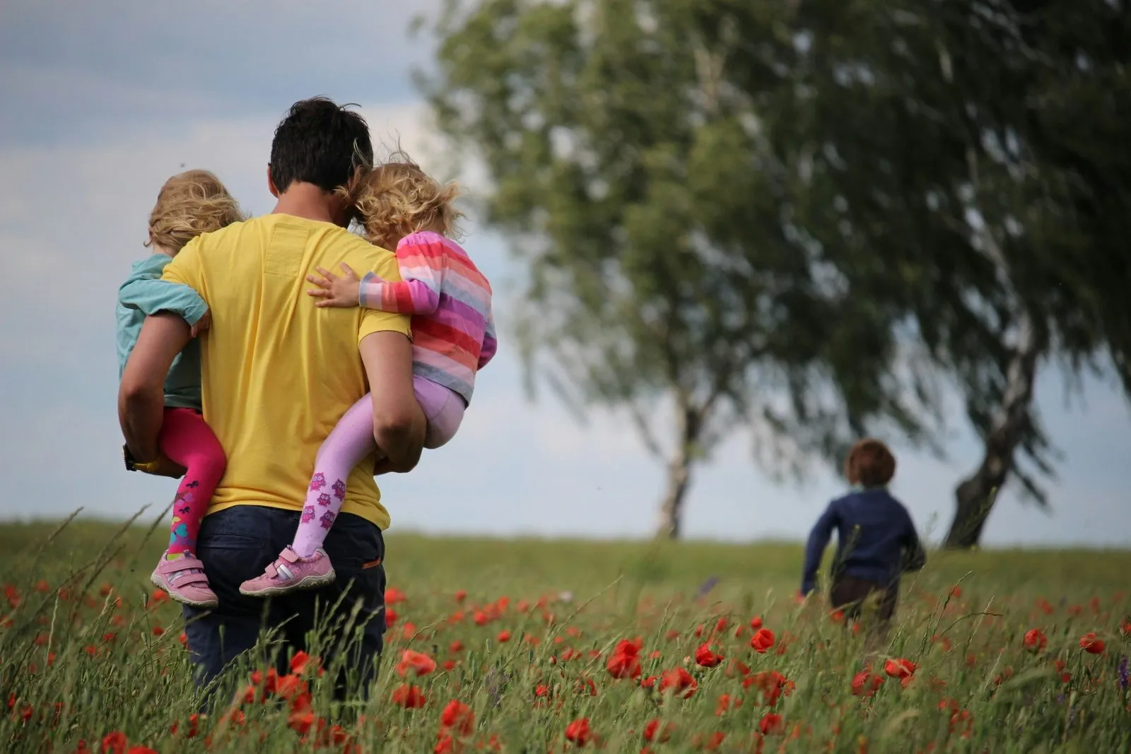 Ein Mann trägt zwei Kleinkinder auf dem Arm durch ein Mohnblumenfeld, ein drittes Kind läuft voraus