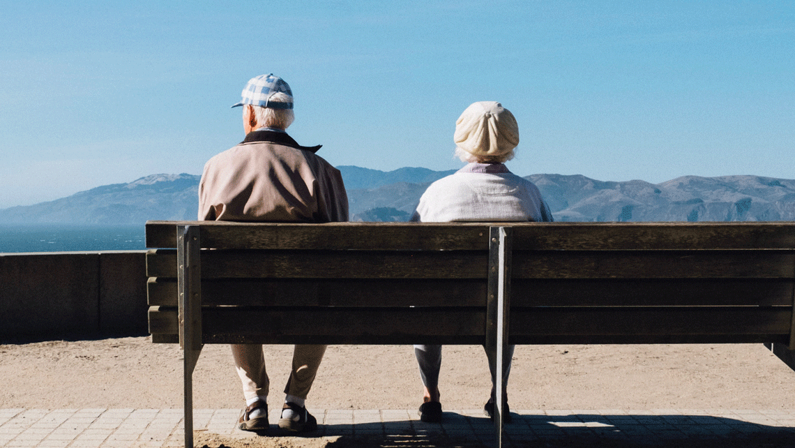 Älteres Paar sitzt von hinten auf einer Bank mit Blick auf Meer und Berge