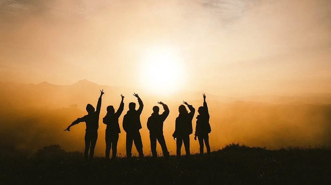 Gruppe jubelt mit erhobenen Armen als Silhouette vor untergehender Sonne