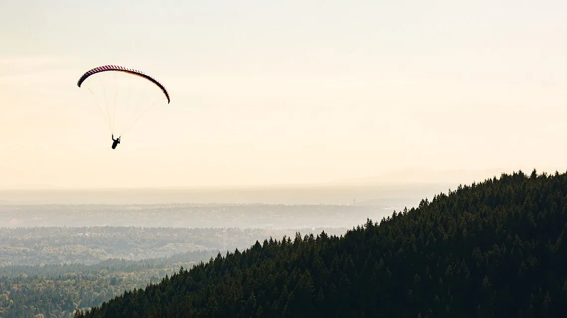 Gleitschirmflieger schwebt über bewaldeten Hügeln im Abendlicht